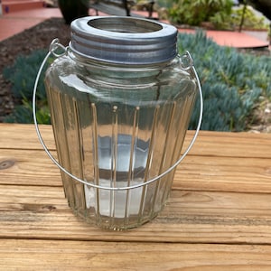 May include: A clear glass jar with a silver lid and a wire handle. The jar has vertical ridges and is sitting on a wooden surface.