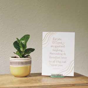 Könnte beinhalten: Eine weiße Karte mit braunem Text und wellenförmigen Linien im Hintergrund. Der Text lautet "Für dich, o Herr, bist du gut und verzeihst Überströmend in standhafter Liebe zu allen, die dich anrufen. Psalm 56:3". Die Karte steht auf einer Holzoberfläche neben einer kleinen grünen Pflanze in einem gelben Topf.