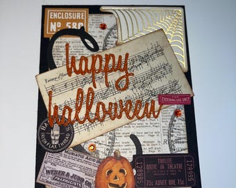 May include: A Halloween card with a black background and a variety of vintage-style embellishments, including a pumpkin, a spiderweb, and a ticket. The card features the text "happy halloween" in orange glitter.