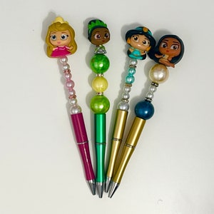 May include: Four colorful pens with princess character toppers and beaded accents. The pens are pink, green, gold, and blue.