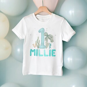 May include: White t-shirt featuring a blue number one, a green sea turtle, and the name "Millie" in blue. The design is surrounded by seaweed and bubbles.