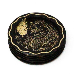 May include: A stack of round black coasters with scalloped edges, featuring gold-colored illustrations of a ship, a flower, and text that reads Cape Cod, Massachusetts, and Cape Cod Bay, with additional small printed details across the surface.