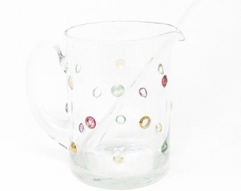 May include: A clear glass pitcher featuring a handle and a small pouring spout, decorated with small, scattered, multi-colored circular accents in shades of pink, yellow, green, and orange. A clear glass stirring rod is placed inside the pitcher.