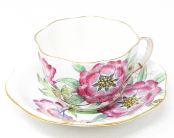 May include: A porcelain teacup and saucer set featuring a white base with a vibrant floral pattern of pink and red flowers with green leaves. The edges of both the cup and the saucer are scalloped and trimmed with a thin gold metallic line.