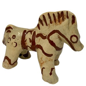 May include: A ceramic figurine of a horse with a cream-colored glaze and hand-painted dark brown markings. The horse features a stylized mane, a saddle-like pattern on its back, and circular details on its hindquarters, all in a glossy finish.