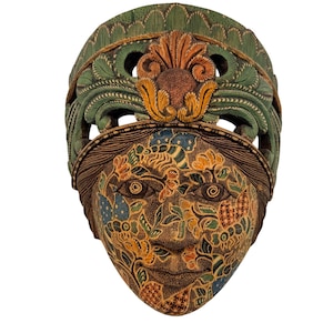 May include: A carved wooden mask featuring a human face adorned with intricate floral and leaf patterns in shades of orange, blue, and green. The mask is topped with a decorative green and orange arched headdress with detailed carved textures.
