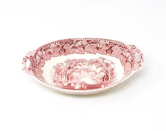 May include: An oval ceramic serving dish featuring a red and white transferware pattern. The rim is decorated with a detailed grape vine motif, while the center depicts a landscape scene with figures, trees, and architectural structures.