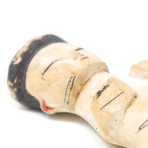 以下が含まれることがあります： 黒髪で目を閉じ、赤い口をした人の小さな手描きセラミック人形。人形は横向きに寝ていて、腕を組んでいます。