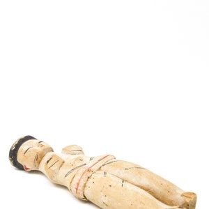 以下が含まれることがあります： 横たわっている人の木製の置物。置物は茶色、白、赤の色合いで塗られています。人は黒い帽子をかぶり、白と赤の衣服を着ています。