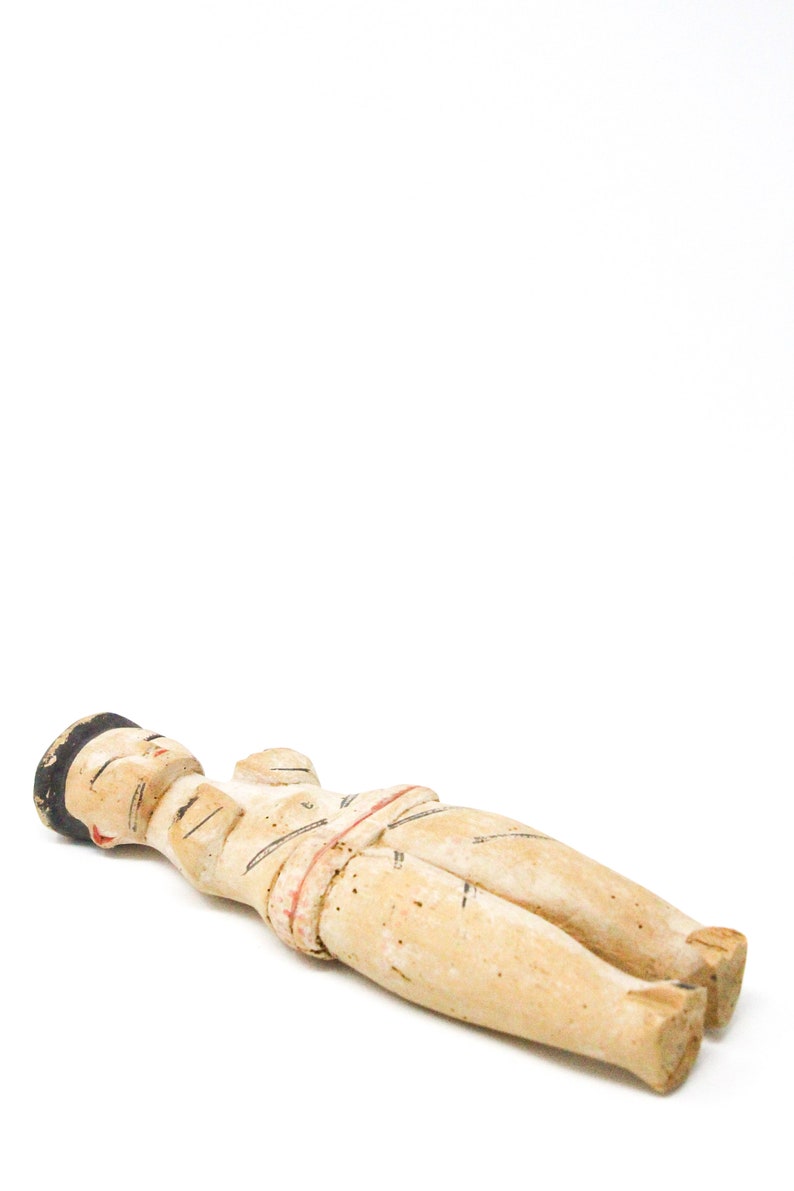 以下が含まれることがあります： 横たわっている人の木製の置物。置物は茶色、白、赤の色合いで塗られています。人は黒い帽子をかぶり、白と赤の衣服を着ています。