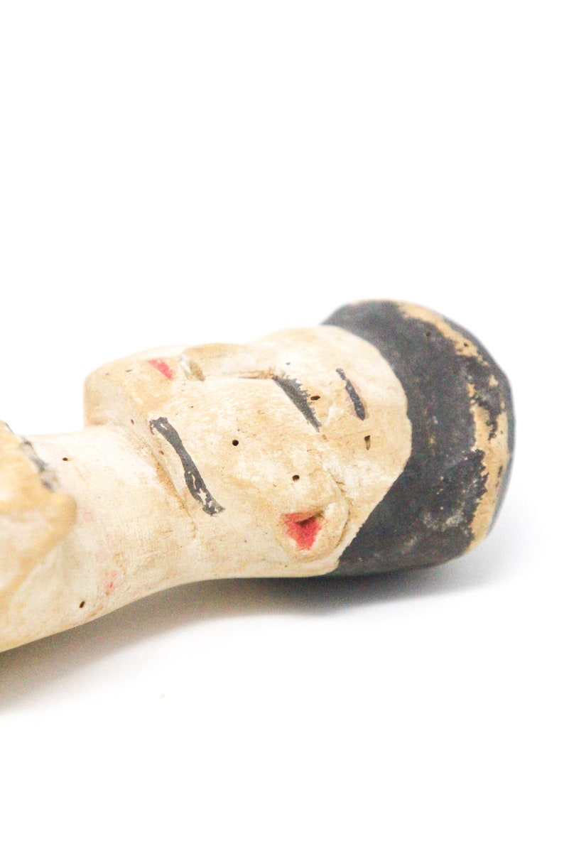 以下が含まれることがあります： 黒髪で目を閉じ、赤い唇をした人の手描きセラミック人形。人形は薄い茶色の粘土で作られており、素朴で手作り感のある外観です。