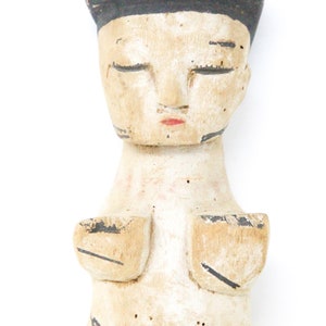 以下が含まれることがあります： 黒い帽子をかぶった人物の、目を閉じた木製の置物。顔は描かれており、置物は白く塗られ、黒と赤のディテールが施されている。