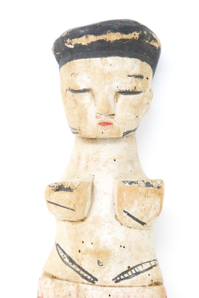 以下が含まれることがあります： 黒い帽子をかぶった人物の、目を閉じた木製の置物。顔は描かれており、置物は白く塗られ、黒と赤のディテールが施されている。