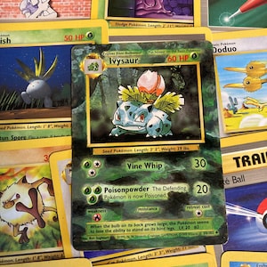 May include: A green, brown and white Pokemon trading card featuring Ivysaur, a Stage 1 Pokemon that evolves from Bulbasaur. The card has a green background with a picture of Ivysaur in the centre. The card text includes the Pokemon's name, type, HP, attacks, weakness, resistance and retreat cost. The card also includes the text "Put Ivysaur on the Basic Pokemon".