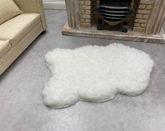 May include: A small, white, cloud-shaped rug made of a fluffy material. The rug is placed on a gray surface, with a miniature sofa and fireplace in the background. This rug is perfect for a dollhouse or miniature scene.