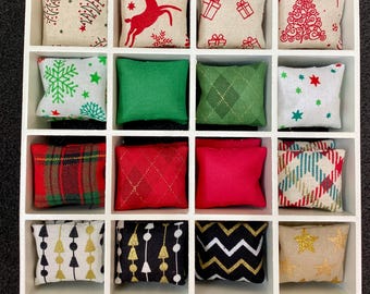 May include: A white box with 16 small, square, fabric pillows. The pillows have various Christmas-themed patterns, including snowflakes, reindeer, plaid, stars, and trees. The pillows are arranged in a grid pattern.