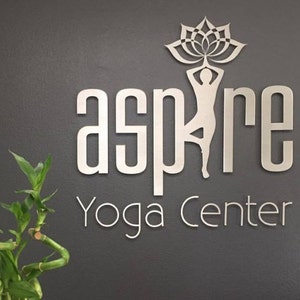 Può includere: Un cartello argentato con la parola "aspire" in grandi lettere e le parole "Yoga Center" in lettere più piccole sotto. Un'immagine stilizzata di una persona che tiene un fiore di loto è sopra la parola "aspire". Una pianta verde si trova nell'angolo in basso a sinistra dell'immagine.
