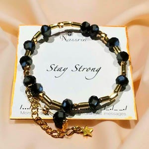 Può includere: Un braccialetto con perline dorate e nere con un ciondolo a forma di stella. Il braccialetto ha le parole "Stay Strong" su un cartoncino bianco.