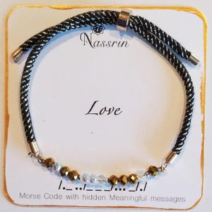 May include: Black and silver braided bracelet with gold beads and clear crystals. The bracelet has a sliding knot closure and the word "Love" is printed on the card. The card also says "Morse Code with hidden Meaningful messages" and shows the Morse code for "Love".