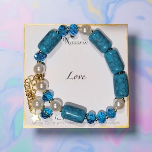 Op de afbeelding: Een turquoise en witte kralen armband met een gouden ster bedel. De armband ligt op een witte kaart met de woorden "Love" en "Nassrin" erop gedrukt. De kaart vermeldt ook "Morse Code met verborgen betekenisvolle berichten".