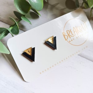 May include: A pair of black and gold triangle stud earrings. The earrings are made of resin and have a geometric design. The earrings are displayed on a white card with the text "Breakout Art Studio" and "breakoutartstudios.com".