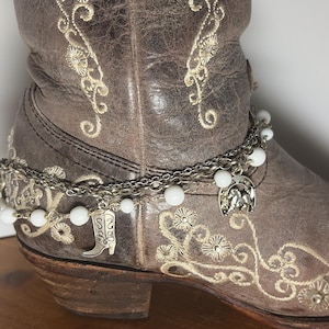 Op de afbeelding: Een bruine leren cowboylaars met ingewikkelde bloemenborduursels en een zilveren enkelbandje. De armband heeft witte kralen en bedels, waaronder een hoefijzer en een cowboylaars.