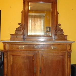 May include: An antique wooden vanity with a large ornate mirror and a marble top. The vanity has two doors and a drawer. The mirror has a carved frame with a fan-shaped design.