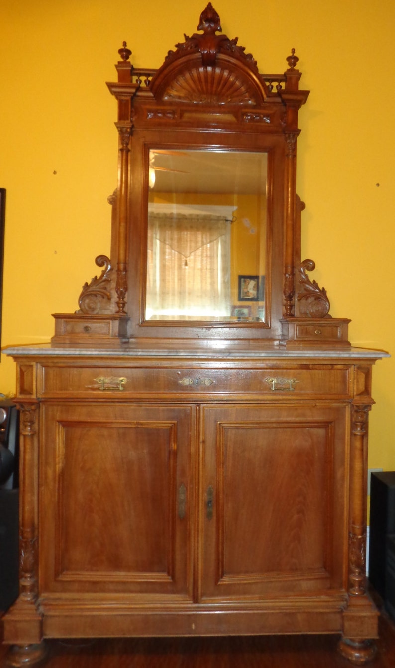 May include: An antique wooden vanity with a large ornate mirror and a marble top. The vanity has two doors and a drawer. The mirror has a carved frame with a fan-shaped design.