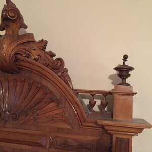 May include: A close-up of the ornate, carved wooden headboard of a bed. The headboard features a large, fan-shaped design in the center, surrounded by intricate scrollwork and floral motifs. The headboard is finished in a rich, dark brown color.