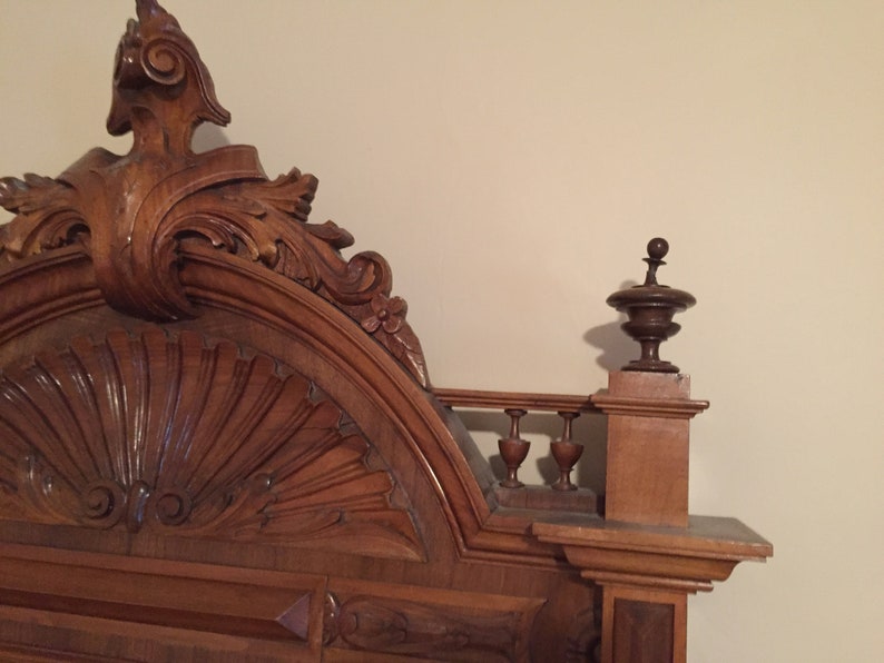 May include: A close-up of the ornate, carved wooden headboard of a bed. The headboard features a large, fan-shaped design in the center, surrounded by intricate scrollwork and floral motifs. The headboard is finished in a rich, dark brown color.