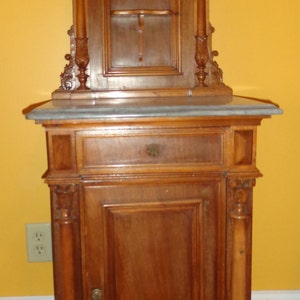 May include: A tall, ornately carved wooden cabinet with a marble top. The cabinet has a single door and a drawer below. The top section has a glass door and a shelf inside.