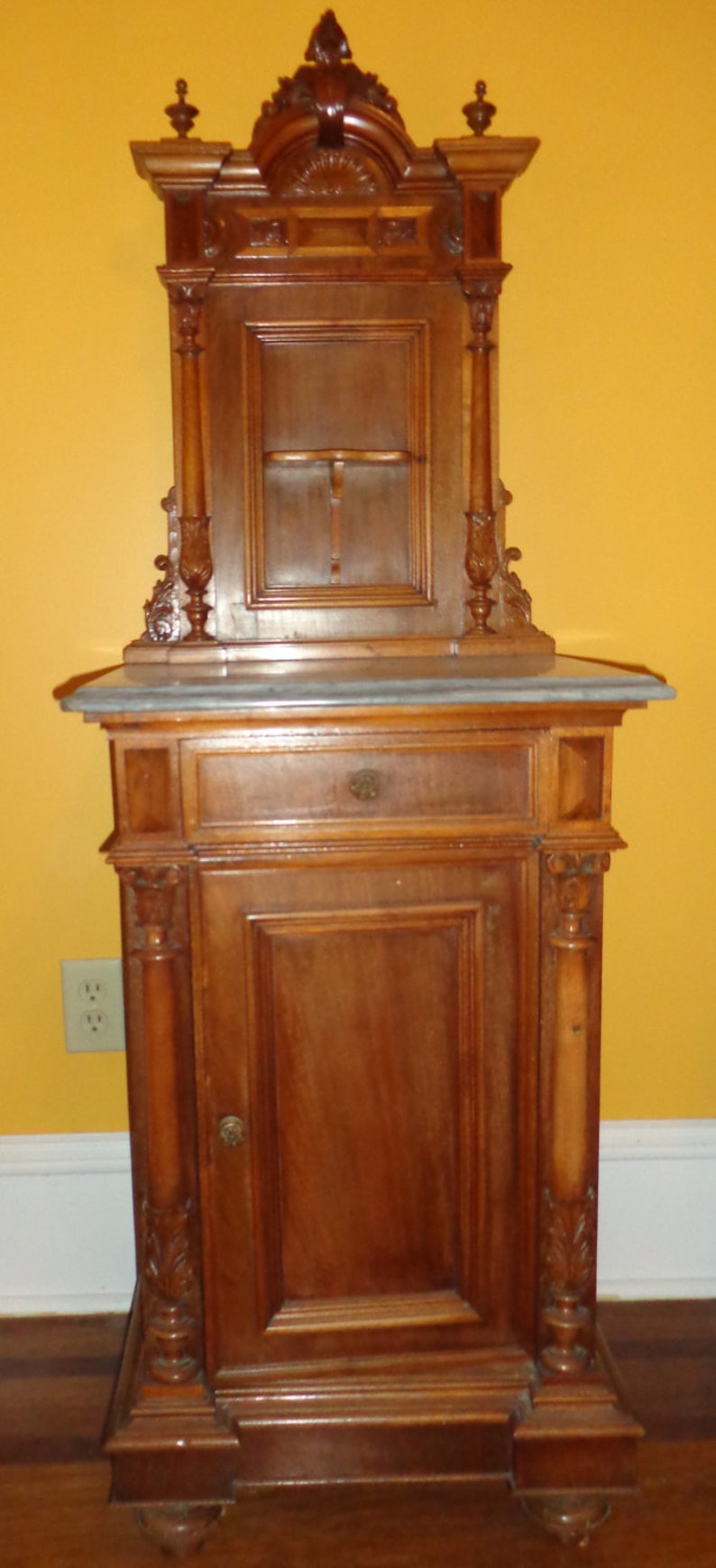 May include: A tall, ornately carved wooden cabinet with a marble top. The cabinet has a single door and a drawer below. The top section has a glass door and a shelf inside.