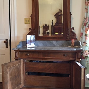 May include: A vintage wooden dresser with a marble top and two drawers. The dresser has two doors that are open, revealing the interior shelves. The dresser is in front of a mirror with a carved wooden frame.