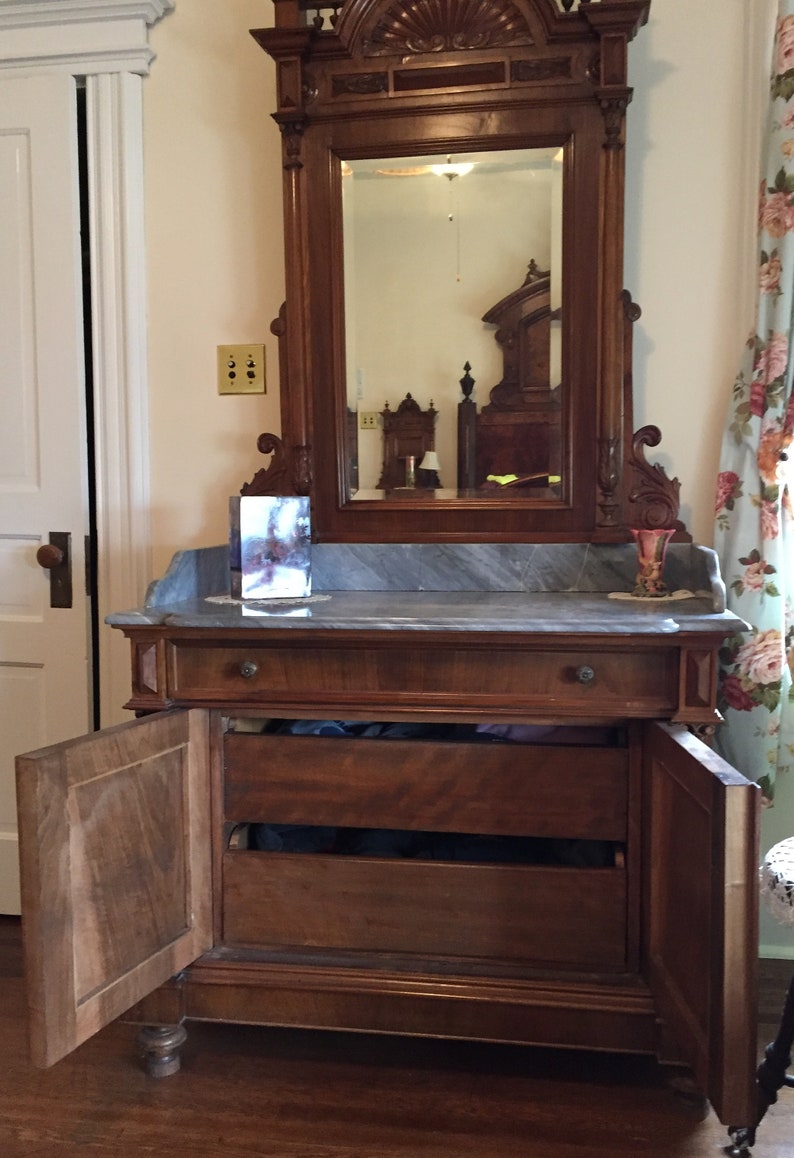 May include: A vintage wooden dresser with a marble top and two drawers. The dresser has two doors that are open, revealing the interior shelves. The dresser is in front of a mirror with a carved wooden frame.
