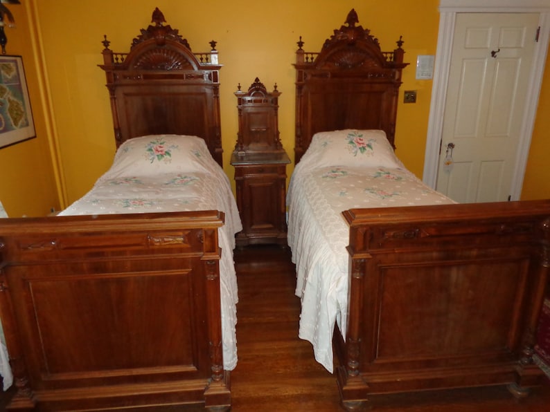 May include: Two antique twin-sized beds with ornate wooden headboards and footboards. The beds are made with dark wood and have white linens with floral embroidery.