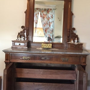 May include: A vintage wooden vanity with a large ornate mirror, marble top, and three drawers. The vanity has a carved wooden frame and a decorative design on the top.