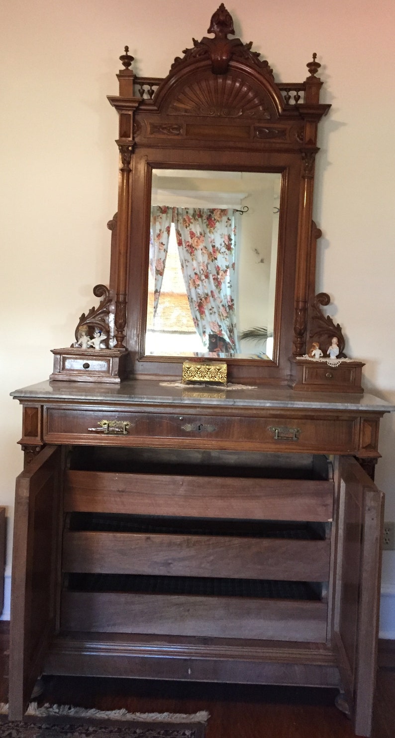 May include: A vintage wooden vanity with a large ornate mirror, marble top, and three drawers. The vanity has a carved wooden frame and a decorative design on the top.