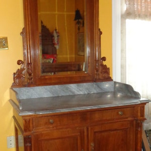 May include: A vintage wooden vanity with a marble top and a large ornate mirror. The vanity has two doors and a drawer, and the mirror is framed with intricate carvings.