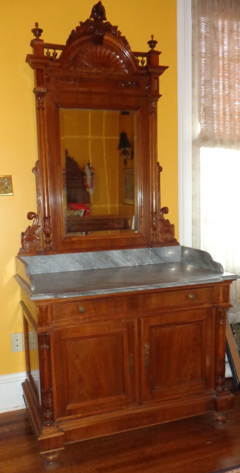 May include: A vintage wooden vanity with a marble top and a large ornate mirror. The vanity has two doors and a drawer, and the mirror is framed with intricate carvings.