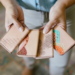 May include: Four tan leather wallets with unique designs are shown. The wallets feature patterns like woven textures, honeycomb, and abstract shapes in brown, orange, and teal. Each wallet has a stitched border, showcasing craftsmanship.