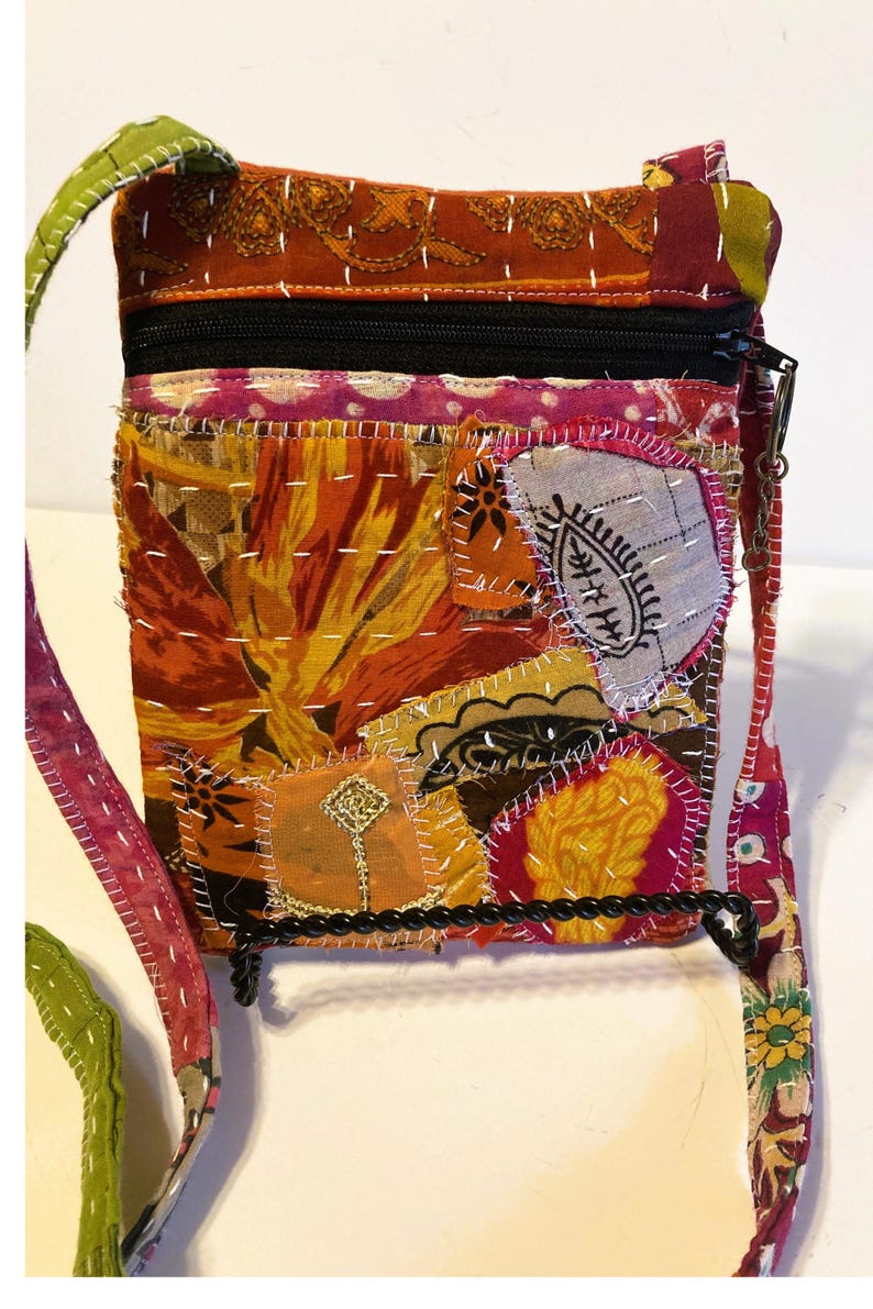 K&ouml;nnte beinhalten: Eine bunte Patchwork-Umh&auml;ngetasche mit schwarzem Rei&szlig;verschluss und Riemen. Die Tasche hat verschiedene Stoffflicken in Orange-, Rot-, Gelb- und Rosat&ouml;nen, die mit wei&szlig;em Faden zusammengen&auml;ht sind. Der Riemen ist eine Kombination aus gr&uuml;nem, rosa und gemustertem Stoff.