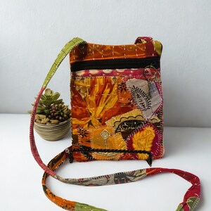 K&ouml;nnte beinhalten: Eine Patchwork-Umh&auml;ngetasche mit farbenfrohem Blumendesign. Die Tasche hat einen schwarzen Rei&szlig;verschluss, einen langen Riemen und besteht aus verschiedenen Stoffflicken in Orange-, Gelb-, Rot- und Gr&uuml;nt&ouml;nen. Die Tasche ist etwa 20 cm hoch und 15 cm breit.