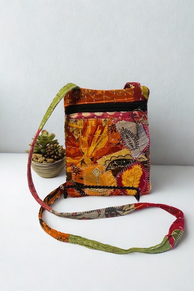 K&ouml;nnte beinhalten: Eine Patchwork-Umh&auml;ngetasche mit farbenfrohem Blumendesign. Die Tasche hat einen schwarzen Rei&szlig;verschluss, einen langen Riemen und besteht aus verschiedenen Stoffflicken in Orange-, Gelb-, Rot- und Gr&uuml;nt&ouml;nen. Die Tasche ist etwa 20 cm hoch und 15 cm breit.