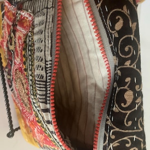 May include: A colorful, patchwork-style bag with a red zipper. The bag features various fabric patterns, including floral and geometric designs in shades of red, yellow, black, and white. The interior lining is striped.