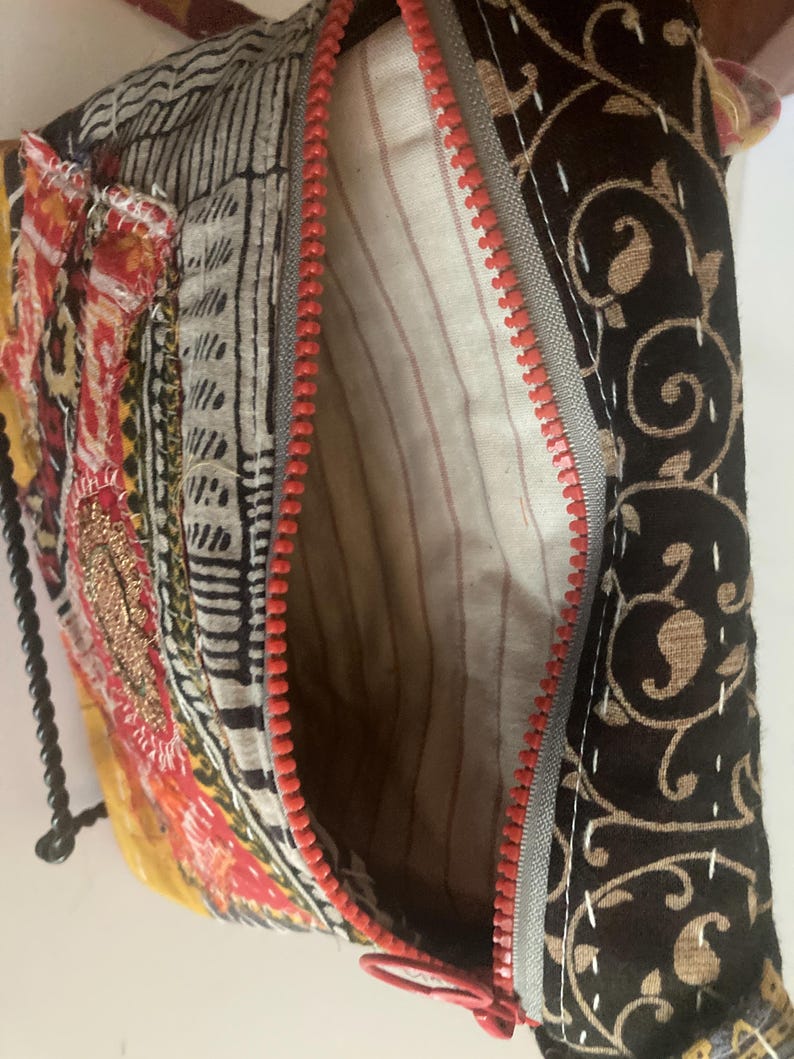 May include: A colorful, patchwork-style bag with a red zipper. The bag features various fabric patterns, including floral and geometric designs in shades of red, yellow, black, and white. The interior lining is striped.