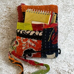 K&ouml;nnte beinhalten: Eine Patchwork-Umh&auml;ngetasche mit verschiedenen Farben und Mustern. Die Tasche enth&auml;lt einen gelben Notizblock und einen roten Stift. Der Gurt ist gr&uuml;n und orange. Die Tasche besteht aus gen&auml;htem Stoff.