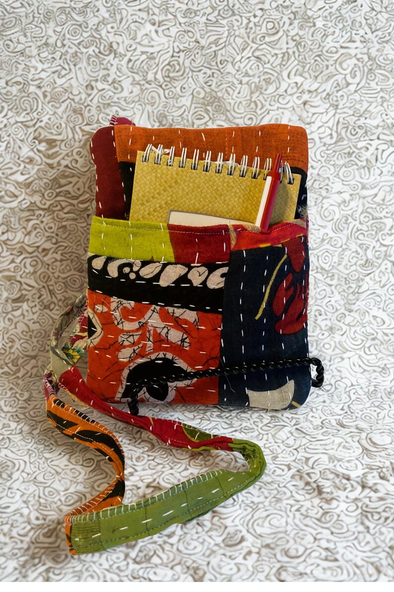 K&ouml;nnte beinhalten: Eine Patchwork-Umh&auml;ngetasche mit verschiedenen Farben und Mustern. Die Tasche enth&auml;lt einen gelben Notizblock und einen roten Stift. Der Gurt ist gr&uuml;n und orange. Die Tasche besteht aus gen&auml;htem Stoff.