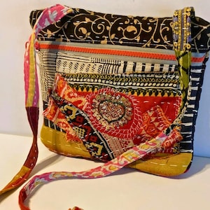 May include: A rectangular crossbody bag constructed from various patches of fabric featuring kantha-style white stitching. The bag includes a bright orange zipper, a central red heart-shaped appliqué with gold sequins, and a long, multicolored strap.