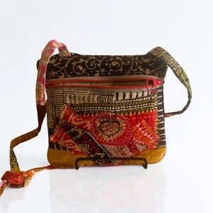May include: A small, square crossbody bag with a patchwork design. The bag features a red zipper, a decorative heart-shaped patch, and a long strap. The color palette includes brown, red, yellow, and patterned fabrics.
