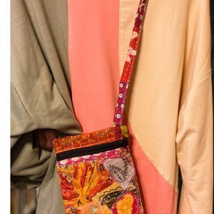 K&ouml;nnte beinhalten: Eine bunte Patchwork-Umh&auml;ngetasche mit floralem Design. Die Tasche hat einen schwarzen Rei&szlig;verschluss und einen gemusterten Riemen. Die Tasche besteht aus verschiedenen Stoffen in Orange-, Rot-, Rosa- und Gelbt&ouml;nen.